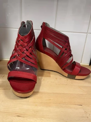 Nya röda kilklackade sandaler från Ecco med flätade remmar - Snygga helt nya röda sandaler från Ecco med kilklack (9,5cm) och flätade remmar i skinnimitation. Skorna har dragkedja bak för enkel påtagning och en stabil träfärgad klack. Perfekta för dig som vill ha både höjd och komfort i sommarens look. Djurfritt och rökfritt hem.