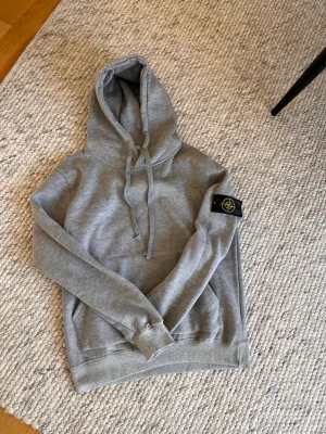 Grå hoodie från Stone Island - Säljer en stilren grå hoodie från Stone Island med klassisk logga på ärmen. Hoodien har dragsko i huvan och en mjuk, skön känsla. Perfekt för dig som gillar streetwear och vill ha något snyggt och bekvämt.