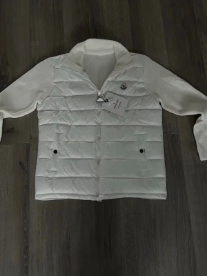 Vit pufferjacka från Moncler - Snygg vit pufferjacka från Moncler med quiltad front och stickade ärmar. Jackan har dragkedja framtill, två fickor med knappar och Moncler-logga på bröstet. Perfekt för kalla vinterdagar och ger en clean, modern look.