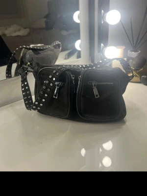 Svart axelväska med nitar och dragkedjor - Cool svart axelväska i mjukt material med två framfickor och silvriga dragkedjor. Väskan har en bred axelrem dekorerad med silvriga nitar för en edgy look. Perfekt för dig som vill ha en praktisk men snygg väska med attityd.