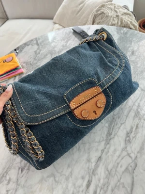 Prada denim handväska med kedjerem - Cool handväska från Prada i blå denim med detaljer i brunt läder och grova kontrastsömmar. Väskan har en kedjerem i metall och läder samt ett lock med snyggt spänne framtill. Perfekt för dig som gillar en avslappnad men lyxig stil. Comes with authentication card from collectors cage and a dust bag, vintage crossbody bag 
