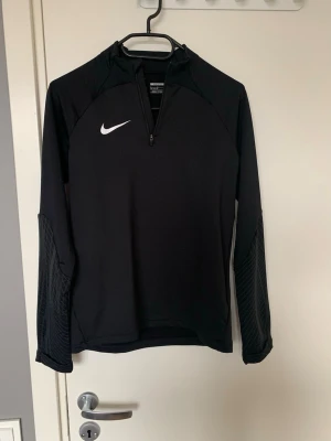 Svart långärmad träningströja Nike - Svart långärmad träningströja från Nike med halv dragkedja framtill och vitt Nike-logga på bröstet. Ärmarna har diskret mönstrad struktur och tröjan är tillverkad i ett stretchigt funktionsmaterial som passar perfekt för träning men också vanligt bruk vardagligen.