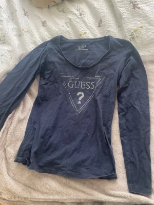 Mörkblå långärmad topp från Guess - Mörkblå långärmad topp från Guess med ikonisk logga i strass framtill. Topp i mjuk bomull med rund halsringning och figurnära passform. Perfekt för dig som gillar stilrena och trendiga plagg med en touch av bling. Lite av glittret har försvunnit som bild visar 