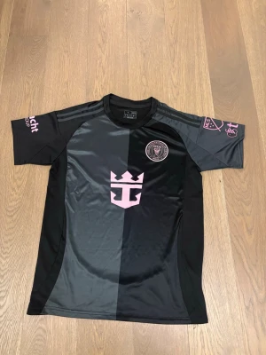 Svart fotbollströja Inter Miami CF - Snygg svart och mörkgrå fotbollströja från Inter Miami CF med rosa detaljer och klubbmärke på bröstet. Tröjan har korta ärmar, slim fit-passform och är tillverkad i ett lätt och ventilerande material. Perfekt för träning eller match.