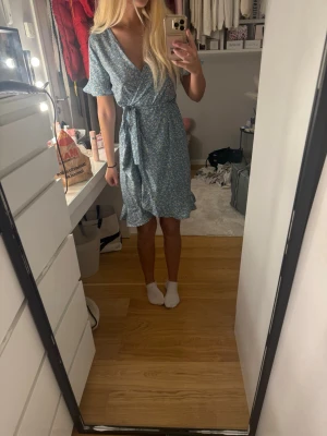 Blommig blå omlottklänning - Söt blå kort omlottklänning med vitt blommönster och volangdetaljer. Klänningen har korta ärmar och knyts i midjan för en smickrande passform. Perfekt för varma dagar och ger en fräsch sommarkänsla.
