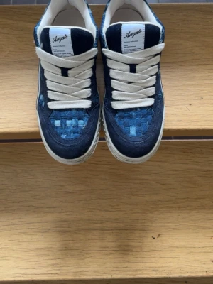 Blåa Angalo sneakers med vävd detalj - Snygga sneakers från Axel arigato i blått med unika vävda detaljer på tån och sidorna. Skorna har vita snören och vit sula, samt en cool broderad logga på sidan. Ovandelen är i en mix av mocka och textil för en modern look. Nypris 4000 mitt pris 2500. Priset kan diskuteras.