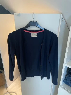 Mörkblå kofta från Holly & Whyte - Stilren mörkblå kofta från Holly & Whyte med rund halsringning och knappar framtill. Koftan har en diskret rosa kant på insidan av kragen och är tillverkad i ett mjukt, tunt material. Perfekt att slänga över en t-shirt eller blus.