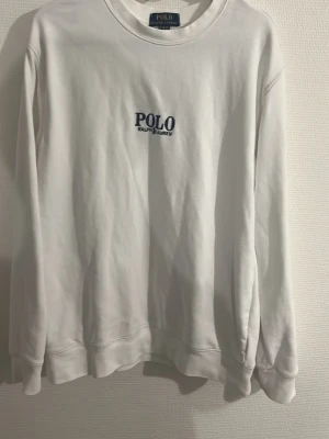 Vit sweatshirt från Polo Ralph Lauren - Vit sweatshirt med rund hals och blå POLO Ralph Lauren-logga broderad på bröstet. Klassisk passform med långa ärmar och ribbade muddar. Perfekt för en clean och stilren look.