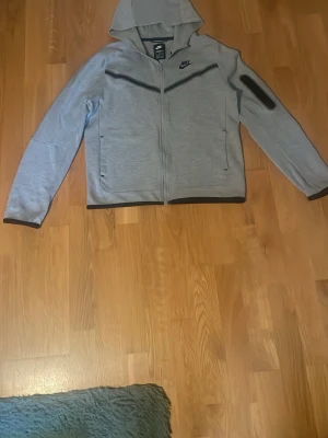 Grå nike tech fleece - Grå hoodie från Nike med hel dragkedja och svarta detaljer längs bröstet. Nike-logga på vänster sida av bröstet. Hoodien har två sidofickor och en extra ficka på ena ärmen. Muddar i svart vid ärmslut och nederkant. Tillverkad i mjukt material för en sportig och bekväm stil.