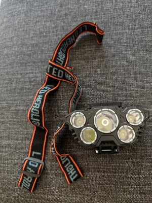 Svart LED-pannlampa med fem lampor - Säljer en svart LED-pannlampa med fem kraftfulla lampor och justerbart elastiskt band med orange kant. Perfekt för utomhusaktiviteter som löpning, camping eller cykling. Bandet har texten 'LED HEADLIGHT' i vitt och lampan ger starkt vitt ljus.