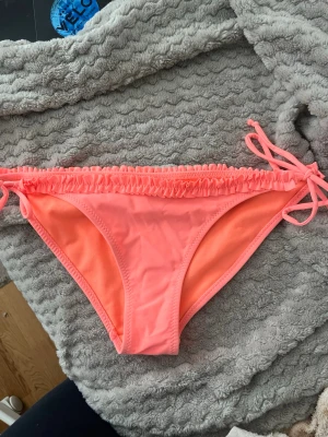 Söt korall bikini  - Snygg rosa bikiniunderdel med knyt i sidorna och rynkade detaljer upptill. Perfekt för sommaren och strandhäng. Tillverkad i mjukt syntetmaterial som torkar snabbt och sitter skönt.
