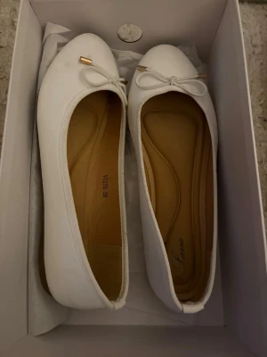 Ballerinaskor  - Säljer ett par vita ballerinaskor med söt rosett och guldfärgade detaljer framtill. Skorna har en mjuk innersula så de är sköna att gå i, använda fåtal gånger 