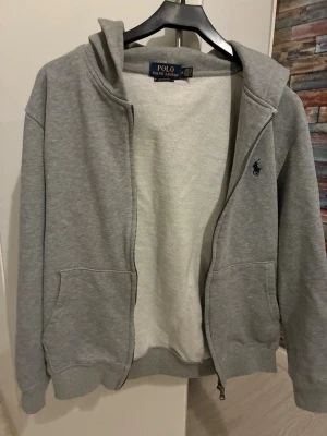 Grå hoodie från Polo Ralph Lauren - Klassisk grå hoodie med dragkedja från Polo Ralph Lauren. Mjuk bomullsblandning, två fickor fram och broderad marinblå logga på bröstet. Perfekt för en avslappnad och stilren look.