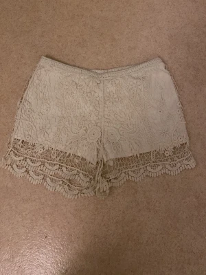 Vita virkade spets-shorts - Säljer ett par vita virkade shorts med vacker spetsdetalj och blommigt mönster. Shortsen har hög midja och en lös passform, perfekta för sommardagar. De har fina volangkanter längst ner!