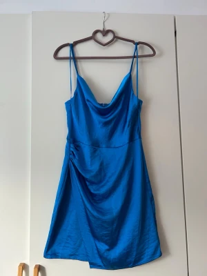 Blå kort klänning med tunna band - Snygg blå kort klänning med tunna axelband och draperad urringning. Klänningen har en lätt glansig finish och en smickrande passform som faller fint. Perfekt för dig som vill ha en enkel men stilren look.