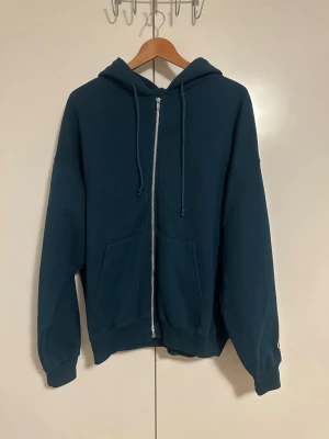 Mörkgrön vintage zip hoodie från Champion - Säljer en mörkgrön hoodie från Champion med dragkedja framtill och två stora fickor. Klassisk huva med dragsko och ribbade muddar vid ärmar och nederkant. Perfekt för en chill och sportig look. Tillverkad i mjukt sweatshirtmaterial.