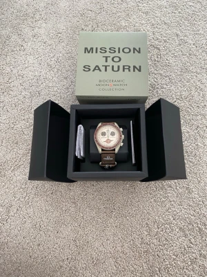 Omega x Swatch Mission to Saturn - Säljer en Omega x Swatch Speedmaster MoonSwatch Mission to Saturn. Klockan har en beige boett i biokeramik, brun bezel och brunt textilband med Omega-logga. Urtavlan är beige med bruna detaljer och Saturnus-motiv. Kommer i originalbox med manual.