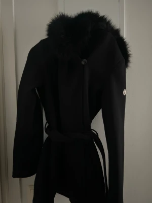 Svart kappa med fuskpäls och bälte - Stilren svart kappa från hollies med stor luva kantad av mjuk fuskpäls. Kappan har ett knytbälte i midjan och en dekorativ metallogo på ärmen. Använd någon gång i några timmar annars helt ny och fin då den inte kommer till användning vill jag sälja den. 