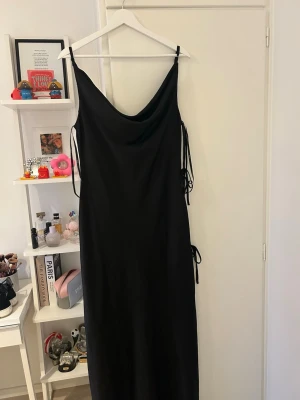 Svart balklänning från ASOS DESIGN - Elegant svart balklänning i satin med slit och tre dekorativa rosetter längs ena sidan som ger en exklusiv och feminin detalj ✨  Perfekt till bal, gala, bröllop eller andra festliga tillfällen 🖤  Klänningen har ett vackert fall och känns lyxig i materialet. Klänningen är helt ny och orginalförpackning medföljer.  Jag är 178 cm lång och bär vanligtvis M och den går nästan ned till fotknölarna, perfekt längd för klackar 👠 