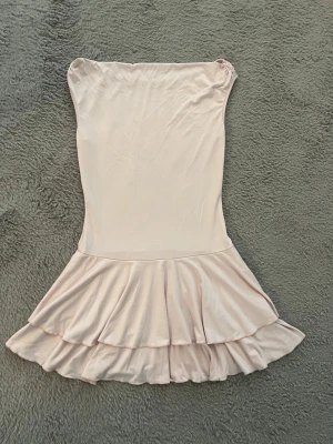 Ljusrosa axelbandslös volangklänning - Söt ljusrosa axelbandslös klänning med volangkant nedtill. Klänningen har en figurnära passform upptill och är sydd i ett mjukt, glansigt material som ger en elegant känsla. Perfekt för dig som gillar en feminin och trendig stil.