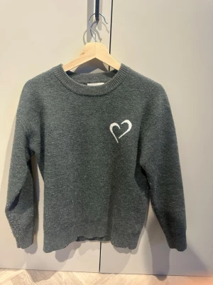 Mörkgrå stickad tröja med hjärta - Mysig mörkgrå stickad tröja med ett vitt broderat hjärta på bröstet. Tröjan har rund halsringning, långa ärmar och ribbade muddar. Perfekt för dig som gillar enkel men söt design. Materialet känns mjukt och värmande.