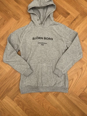 Grå hoodie från Björn Borg 170cm - Snygg grå hoodie från Björn Borg med svart text på bröstet där det står 'BJÖRN BORG STOCKHOLM 1956'. Hoodien har huva med dragsko, magficka och ribbade muddar vid ärmar och nederkant. Passar dig som är ungefär 170cm (XS-S) Tillverkad i mjukt sweatshirtmaterial, perfekt för en avslappnad stil.