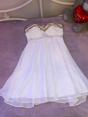 Vit axelbandslös klänning Elise Ryan - Säljer en vit axelbandslös kortklänning från Elise Ryan med vacker guldfärgad detalj upptill. Klänningen har ett lätt och svepande tyg i flera lager och en feminin silhuett. FINNS PÅ FLER SIDOR!!