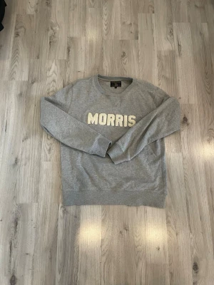 Grå sweatshirt från Morris - Säljer en grå sweatshirt från Morris med stora vita bokstäver broderade på bröstet. Tröjan har rund halsringning, ribbade muddar vid ärmslut och nederkant samt en klassisk relaxed fit. Perfekt för en chill och stilren look.
