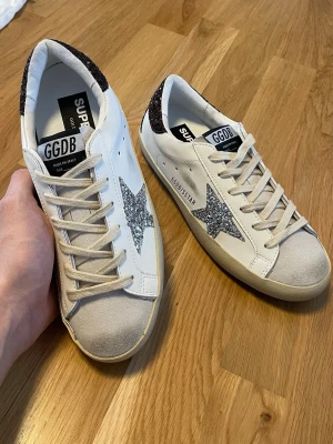 Golden Goose vita sneakers med glitter - Golden Goose sneakers i vitt läder med silverglittrig stjärna på sidan och svartglittrig häl. Klassisk låg modell med beige mockadetaljer på tån och vita snören. Snygg kontrast mellan glitter och läder för en unik look.