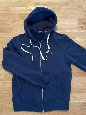 Marinblå hoodie från Polo Ralph Lauren - Snygg marinblå hoodie från Polo Ralph Lauren med dragkedja, vit dragsko och klassisk röd logga på bröstet. Tillverkad i mjuk bomullsblandning och har två fickor framtill samt ribbade muddar. Perfekt för en avslappnad och stilren look.