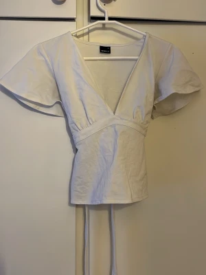 Vit omlottopp med v-ringning - Snygg vit omlottopp från NA-KD med djup v-ringning och vida korta ärmar. Toppen har knytband i midjan för en justerbar passform och är tillverkad i mjuk bomull. Perfekt för en trendig och avslappnad stil.