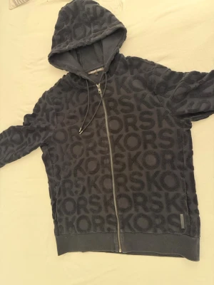 Svart hoodie från Michael Kors - Snygg svart hoodie från Michael Kors med dragkedja och stor huva. Hoodien har ett upphöjt KORS-mönster över hela plagget och ribbade muddar vid ärmslut och nederkant. Tillverkad i mjukt material som känns lyxigt och bekvämt. Perfekt för en trendig och avslappnad stil.