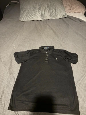 Svart pikétröja från Polo Ralph Lauren - Klassisk svart pikétröja från Polo Ralph Lauren med vit broderad logga på bröstet. Tröjan har korta ärmar, krage och knappar framtill. Perfekt till jeans eller shorts för en clean och stilren look.