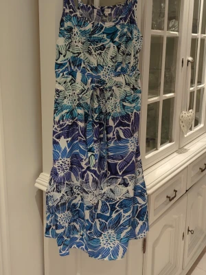 Blommig blå och vit sommarklänning - Säljer en ärmlös klänning med stort blommönster i blått, vitt och turkost. Klänningen har knyt i midjan. Justerbara axelremmar. 