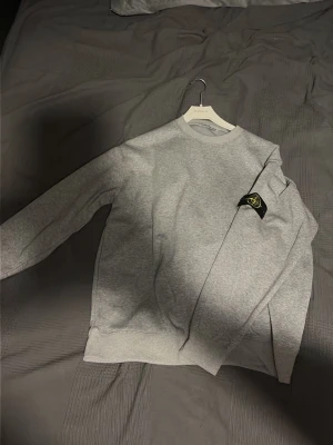 Grå sweatshirt från Stone Island - Säljer en stilren grå sweatshirt från Stone Island med klassisk rund halsringning och ikonisk logotyp-patch på vänster ärm. Tröjan har långa ärmar och är tillverkad i mjukt bomullsmaterial. Perfekt för en avslappnad och trendig look.