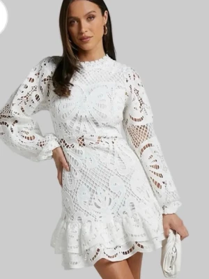 Vit spetsklänning med volang - Supersöt vit kort klänning i spets med långärmade ärmar och volangkant nertill. Klänningen har höghalsad design och fina broderade detaljer i spetsmaterial. Perfekt för dig som vill ha en romantisk och trendig look.