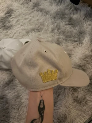 Beige New Era keps med guldkrona - Snygg beige keps från New Era med en glittrig guldkrona broderad på sidan. Smutsig och söndrig brim. Är självklart öppen för prisförslag och säljer fler varor så köper du många får du också mängdrabatt. Obs säljes som på bild.