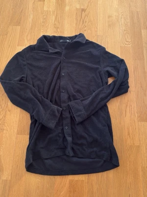 Svart långärmad skjorta i frotté  - En enkel svart skjorta med lång ärm och knappar framtill. Skjortan har klassisk krage och är gjord i mjuk frotté , perfekt för en avslappnad stil. Passformen är normal och tyget känns bekvämt mot huden.