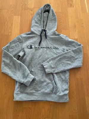 Grå hoodie från Champion - Snygg grå hoodie från Champion med stor logga broderad på bröstet. Klassisk känguruficka framtill och justerbar huva med svarta snören. Tillverkad i mjukt bomullsmaterial som är perfekt för chill dagar eller streetwear-stil.