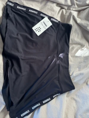 Svart Padelkjol  från Stronger - Säljer en svart Padelkjol från Stronger med logga längs midjan. Shortsen är tillverkade i ett stretchigt och snabbtorkande material, perfekta för träning eller gym. Snygg passform och bekväm midja.