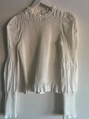Vit smockad blus med puffärm - Trendig vit blus med smockdetaljer över bröstet och ärmsluten. Puffiga långärmar och volangkant nedtill. Perfekt att styla med jeans eller kjol💕💕