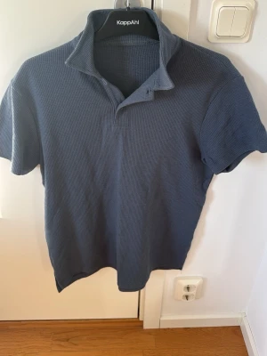 Blått loungset från KappAhl - Mysigt loungset från KappAhl i blått, ribbat material. Setet består av en kortärmad pikétröja med krage och knappar samt matchande shorts med dragsko och fickor. Perfekt för chill hemma eller som sovplagg.