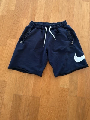 Marinblå Nike shorts med snörning - Snygga marinblå shorts från Nike med vit snörning i midjan och dragkedjeförsedda fickor. Stort vitt Nike-logga på vänster ben. Perfekta för sommarens alla aktiviteter.