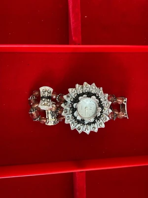 Glittrig silverfärgad JL klocka - Unik klocka från JL med ett stort, blomformat ur i silver och svart, dekorerad med gnistrande stenar. Armbandet består av pärlliknande detaljer och metallspänne. Perfekt för dig som gillar statement-accessoarer och vill sticka ut.