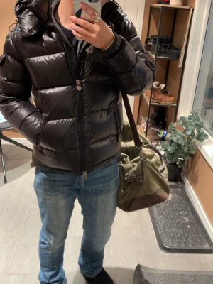 Svart dunjacka från Moncler - Snygg svart dunjacka från Moncler med glansig finish och quiltad design. Jackan har huva, dragkedja framtill och två sidofickor. Perfekt för kalla vinterdagar och ger en riktigt clean look. Klassisk Moncler-logga på insidan.