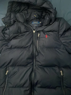  Äkta Svart pufferjacka från Polo Ralph Lauren - Snygg svart pufferjacka från Polo Ralph Lauren med huva och dragkedja framtill. Jackan har två sidofickor med dragkedja och den klassiska röda loggan broderad på bröstet. Perfekt för kalla vinterdagar och riktigt stilren.