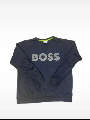 Mörkblå sweatshirt från BOSS - Mörkblå sweatshirt från BOSS med stort vitt logotryck på bröstet. Tröjan har rund halsringning, ribbade muddar vid ärmslut och nederkant samt en grön detalj på insidan av kragen. Perfekt för en avslappnad och stilren look.
