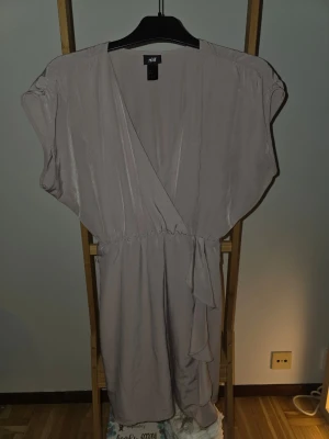 Beige omlottklänning från H&M - Säljer en beige omlottklänning från H&M med korta ärmar och knyt i midjan. Klänningen har en v-ringad design och är tillverkad i ett mjukt, lätt material som ger ett snyggt fall. Perfekt för dig som gillar enkel men stilren look.