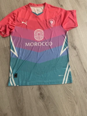 Färgglad Morocco fotbollströja Puma - Säljer en färgstark fotbollströja från Puma med MOROCCO-tryck. Tröjan har rosa, lila och turkosa partier i geometriskt mönster, korta ärmar och DryCell-material som andas. Puma-logga på bröstet och emblem på vänster sida. Perfekt för träning eller match!