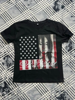 Svart t-shirt med USA-flagga och tryck - Cool svart t-shirt med tryck av amerikanska flaggan och ett konstnärligt motiv av ett ansikte på framsidan. T-shirten har rund hals och korta ärmar. Tillverkad i mjuk bomull, perfekt för en avslappnad streetwear-look.